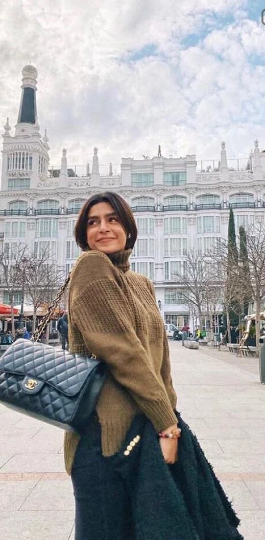 Angely in an olive sweater with a black bag, standing in a city plaza